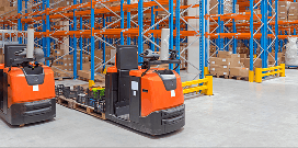 Electric pallet jacks parked in a warehouse aisle with tall storage racks and boxed inventory in the background.