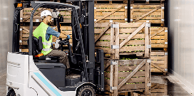 A worker uses a counterbalanced forklift to pick up two crates in front of a stack of other crates