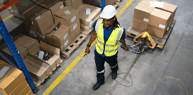 Warehouse worker wearing a hard hat and safety vest walking through an aisle with a pallet jack.