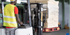 Forklift operator moving stacked materials inside a warehouse while wearing a safety vest and hard hat.