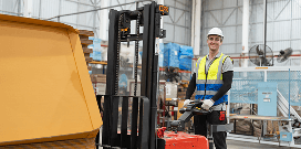 Warehouse worker operating an electric pallet stacker while wearing a hard hat and safety vest.