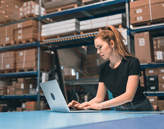 A worker uses a laptop on top of a large table in front of a shelving unit holding inventory
