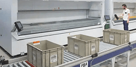 Plastic bins moving along a conveyor system in an automated warehouse with a worker monitoring operations nearby.