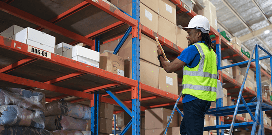 A worker wearing safety attire uses a work platform to inspect packages on a high shelf
