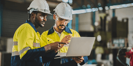 Two industrial workers in safety helmets reviewing information on a laptop inside a manufacturing or warehouse facility.