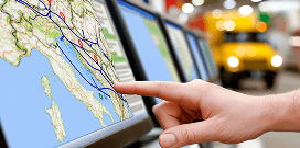 Hand pointing at a digital map on a logistics tracking screen, with a delivery truck blurred in the background.
