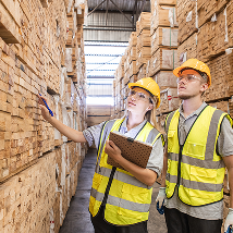Two workers wearing safety attire look at stacks of lumber in a warehouse facility